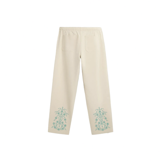 Cream-colored drawstring sweatpants with a relaxed, straight-leg fit. They feature teal ornate floral motifs near the bottom of each leg and a small teal logo on the upper left thigh.