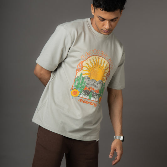 Beige crewneck sweatshirt featuring a retro-style graphic of a large yellow sun over a desert landscape with cacti, mountains, and sunflowers. The word “CALIFORNIA” arcs above the sun, and “dreaming” appears below the illustration.