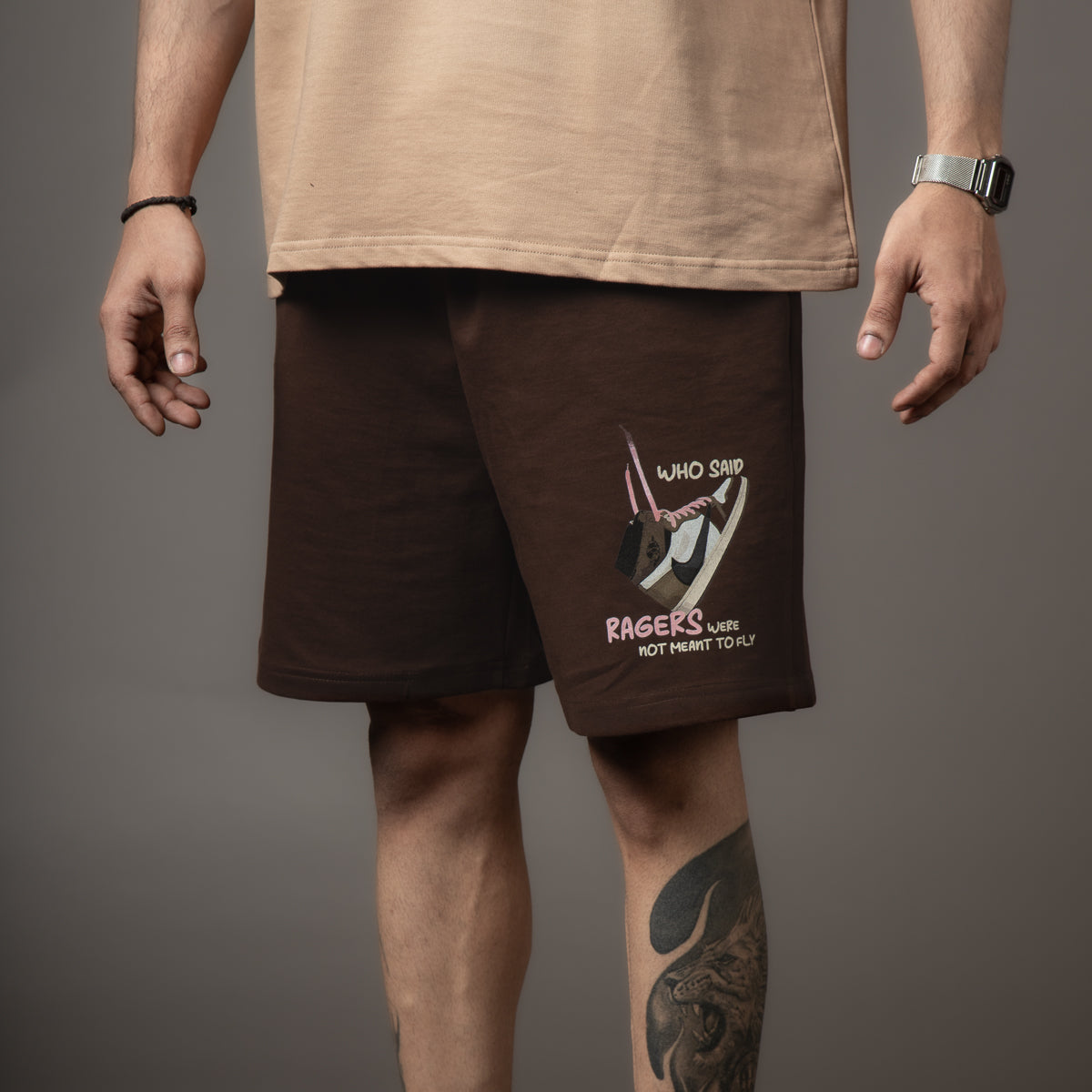 A person wearing brown shorts with a graphic of a sneaker and text "Who said ragers were not meant to fly." Casual vibe, neutral background.