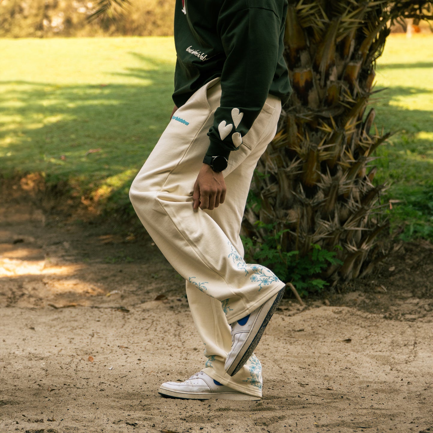 Cream-colored drawstring sweatpants with a relaxed, straight-leg fit. They feature teal ornate floral motifs near the bottom of each leg and a small teal logo on the upper left thigh.