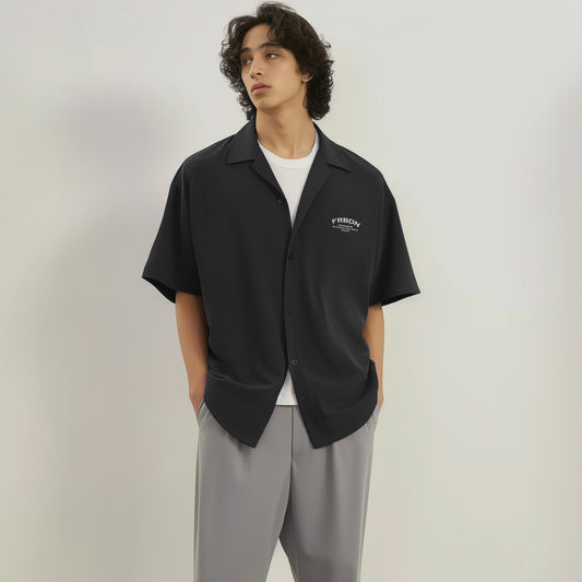 Model wearing a black short-sleeve button-up shirt with a relaxed collar and a small white “FRBDN” graphic on the left chest, paired with grey trousers.