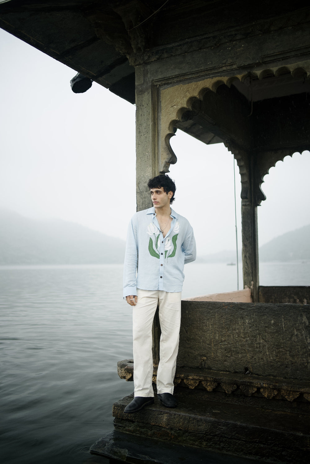 Person wearing light blue full sleeve shirt standing on a old fort structure by a body of water with a misty background