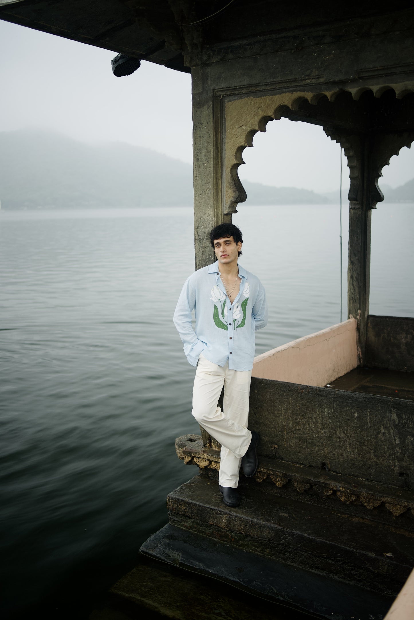 Person wearing light blue full sleeve shirt standing on a old fort structure by a body of water with a misty background