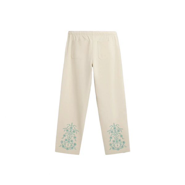 Cream-colored drawstring sweatpants with a relaxed, straight-leg fit. They feature teal ornate floral motifs near the bottom of each leg and a small teal logo on the upper left thigh.