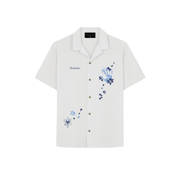 The design includes layered flowers, petals, and small decorative elements in varying shades of blue. The word "Forbidden" is embroidered on the left chest in a small script font. The buttons are metallic or bronze-toned, adding a subtle contrast to the clean white shirt. The overall aesthetic is elegant, artistic, and slightly whimsical.